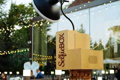 Fotobox aus hellem Holz mit Studioblitz in einem Garten mit Lichterketten in der Dämmerung, im Hintergrund Gäste