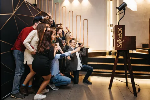 Viele Gäste gehen in die Hocke und rücken zusammen für ein Gruppenfoto vor einer Fotobox in einer warm beleuchteten Halle