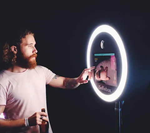Bärtiger Mann im rosa T-Shirt tippt bei einem Abendevent auf den Touchscreen in einem großen weißen Ringlicht