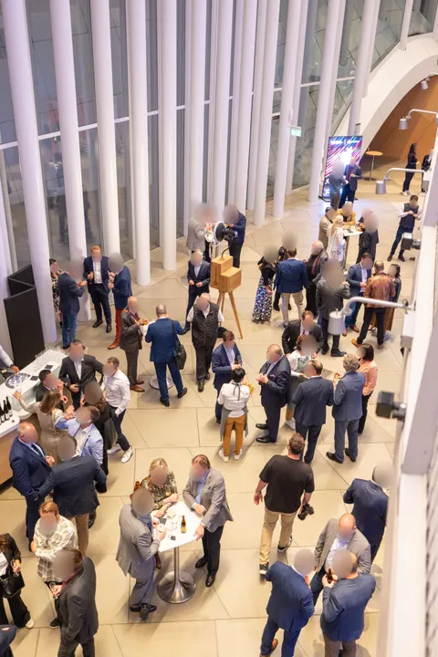 Vue d’en haut : une réception dans un foyer lumineux aux colonnes blanches, des invités en costume autour de mange-debout et l’Instabox W en chêne sur son trépied parmi eux