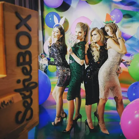Fotobox-Foto: Vier Frauen in Paillettenkleidern posieren mit Partyrequisiten vor einem bunten Hintergrund voller Luftballons