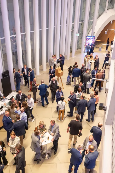 Blick von oben auf einen gut besuchten Empfang: Gäste in Businesskleidung an Stehtischen in einer hellen Halle