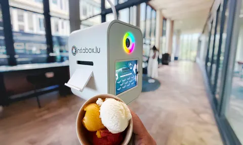 Le photobooth blanc Instabox M et son anneau lumineux arc-en-ciel dans un couloir clair, une coupe de glace tenue au premier plan
