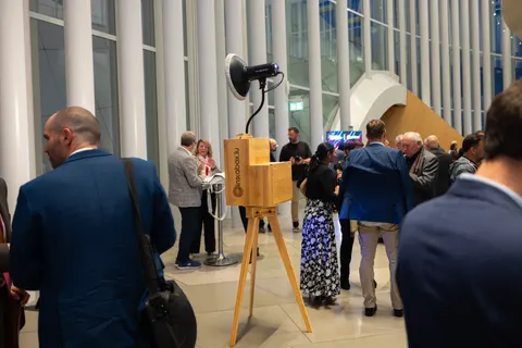 Gäste im Anzug stehen an und plaudern neben einer Fotobox aus Eiche bei einem Abendempfang in einer Säulenhalle