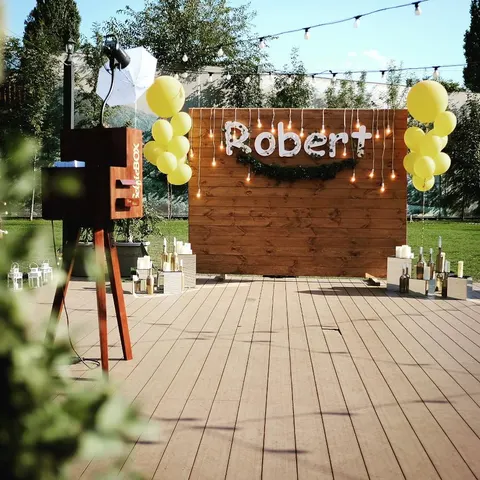 Gartenfest mit einer Holz-Fotobox vor einer Holzwand mit beleuchtetem Namensschild, gelben Ballons und Lichterketten