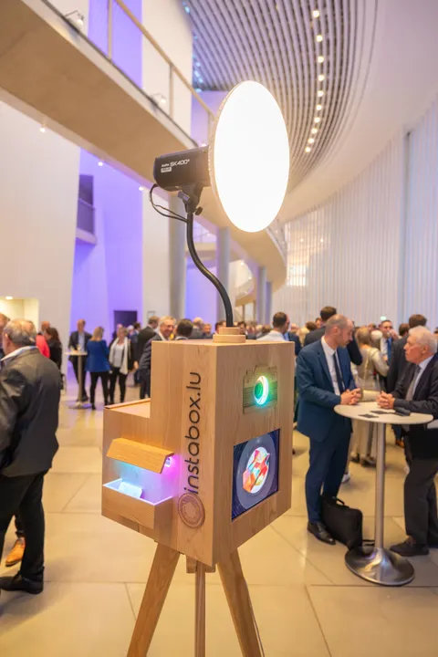 Fotobox aus Holz mit runder Leuchte auf einem Stativ in einem hellen Kongressfoyer, Gäste im Anzug an Stehtischen