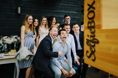 Fotobox-Foto: Freunde ziehen Grimassen vor einer dunklen Holzwand bei einem Empfang, dahinter Frauen in Sommerkleidern