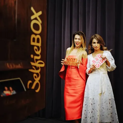 Fotobox-Foto: Zwei Frauen in eleganten Kleidern, korallenrot und weiße Spitze, posieren abends mit lustigen Sprechblasen
