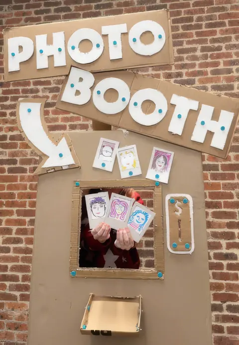 Photobooth en carton aux lettres découpées contre un mur de briques, quelqu’un derrière le trou tient des photos dessinées